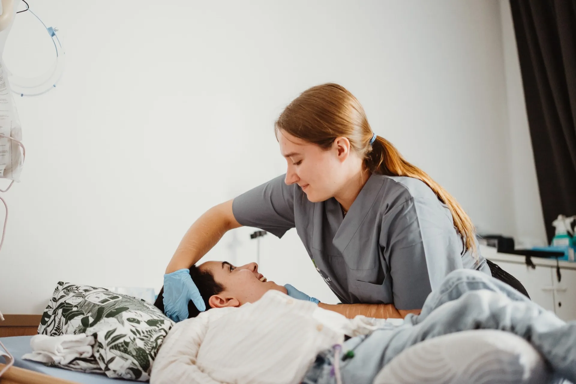 Eine Pflegekraft von Bernstein in grauer Dienstkleidung beugt sich lächelnd über einen jungen Patienten im Pflegebett, daneben ein Infusionsständer.